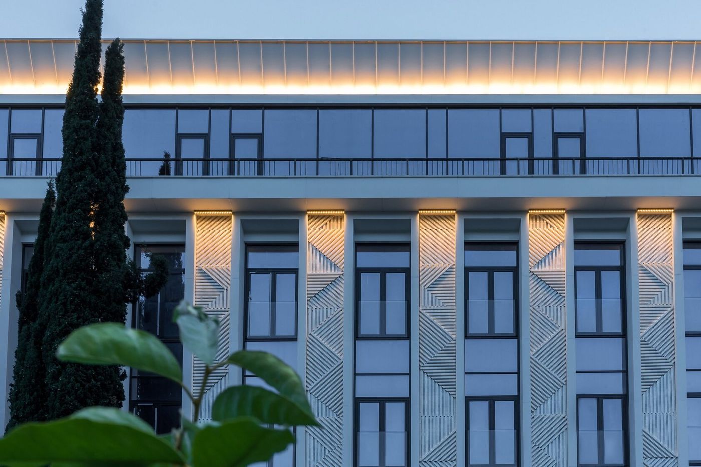 Фасад отеля крупно: белые пилоны с рельефным узором, подсветка под карнизом, верхний этаж с панорамным остеклением, слева кипарис