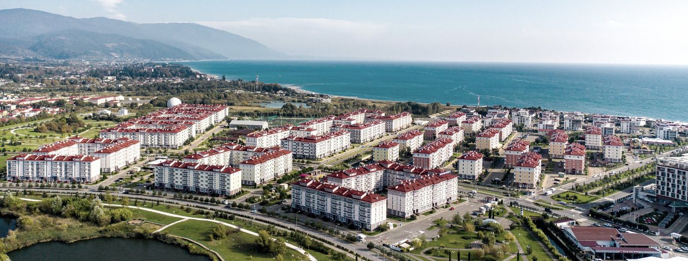 Панорама сверху: кварталы из многоэтажных корпусов с красными крышами, рядом озеро и парк, дальше — побережье и горы