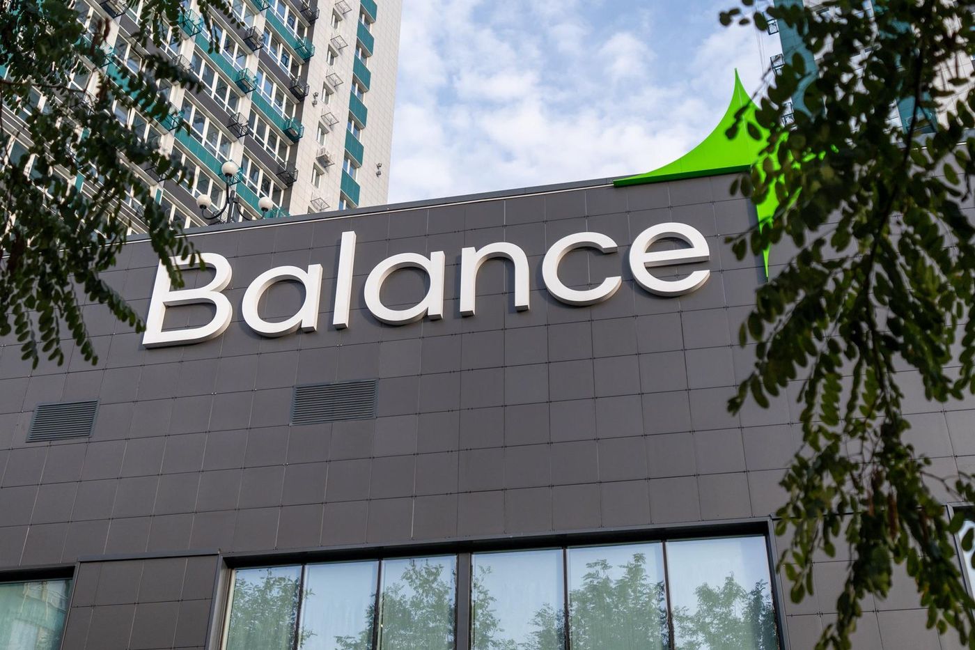Вывеска Balance на тёмном фасаде здания фитнес-клуба, рядом ветви дерева и высотный дом