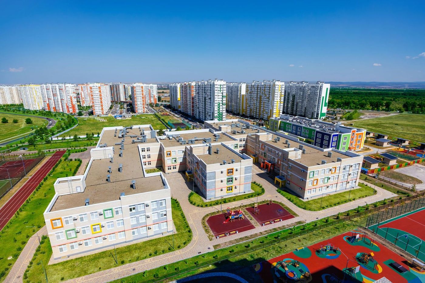 Вид сверху на трёхэтажные корпуса с цветными оконными рамами, спортивное поле с беговой дорожкой и детские площадки