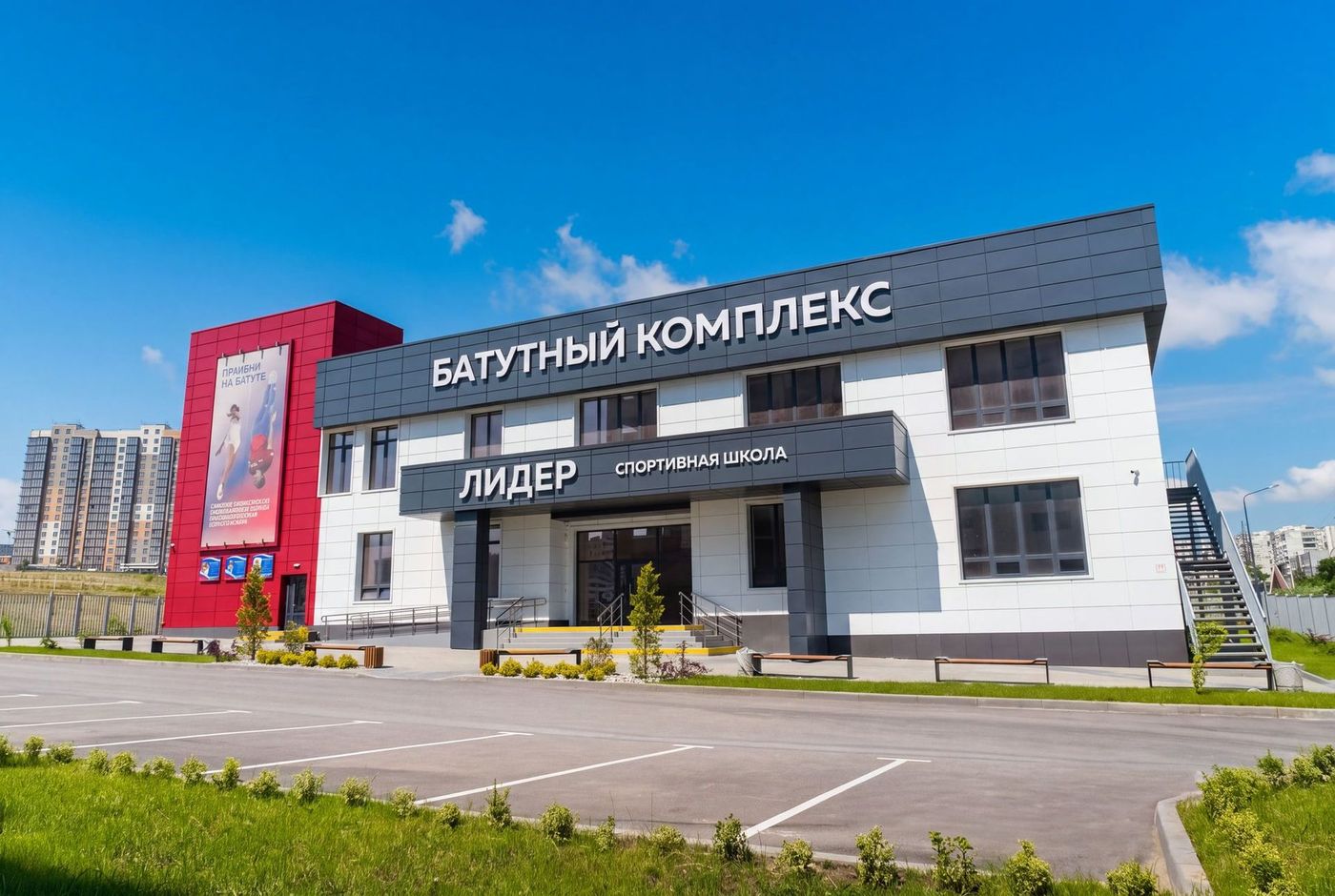 Двухэтажное здание батутного комплекса «Лидер» с красным фасадом у входа и парковкой перед зданием