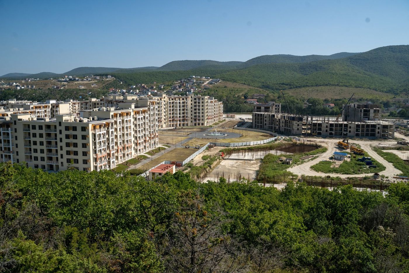Панорама с холма: светлые дома комплекса и стройплощадка в долине, вокруг лесистые холмы и село