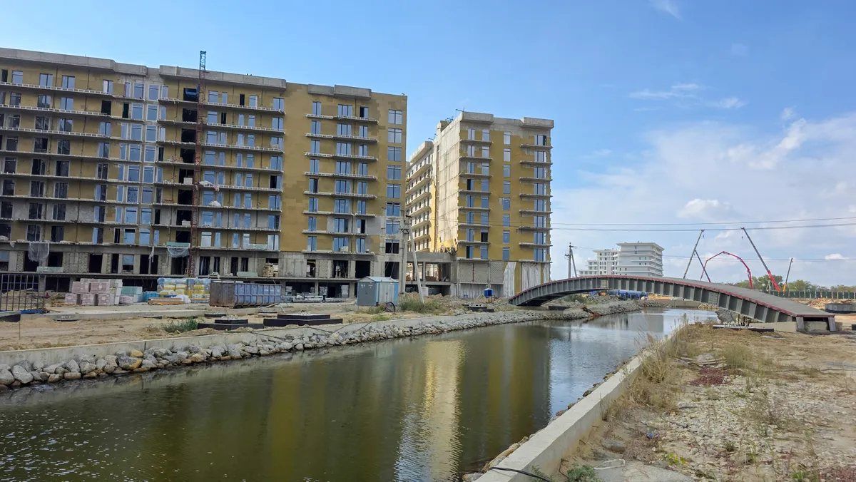 Канал с укреплёнными камнем берегами и пешеходный мост, за ними строящиеся корпуса