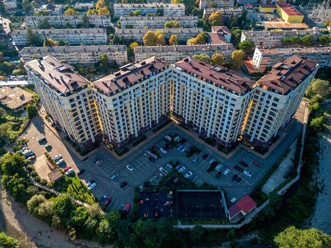 Вид сверху на четыре корпуса с коричневыми крышами, выстроенные ломаной линией, перед ними парковка и спортивная площадка