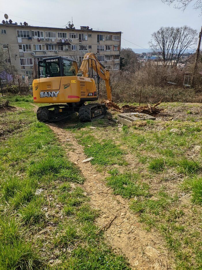 Экскаватор на заросшем травой участке, за ним пятиэтажный дом