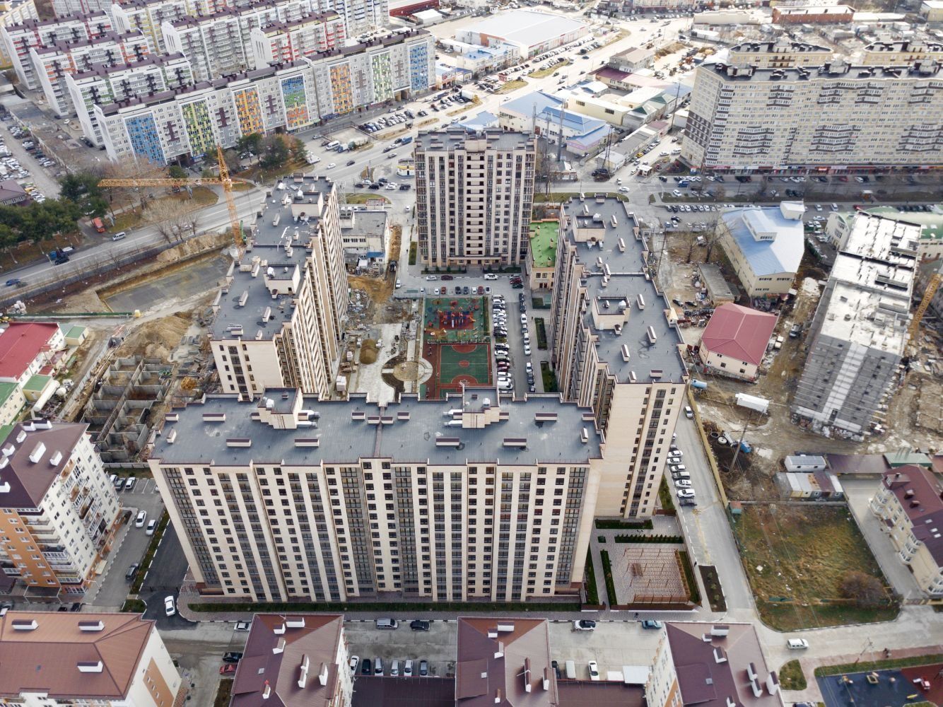 Вид сверху на корпуса комплекса вокруг двора со спортивной площадкой, вокруг городская застройка и стройки