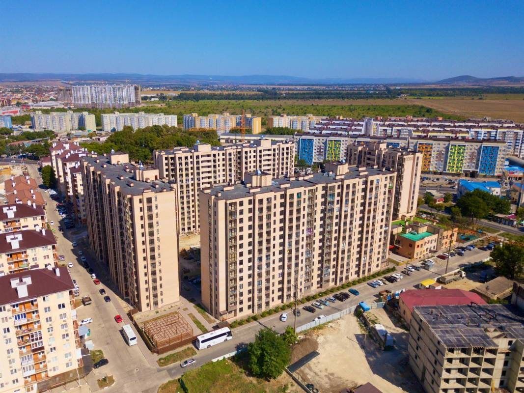 Корпуса комплекса сверху среди городской застройки, дальше поля и холмы под ясным небом