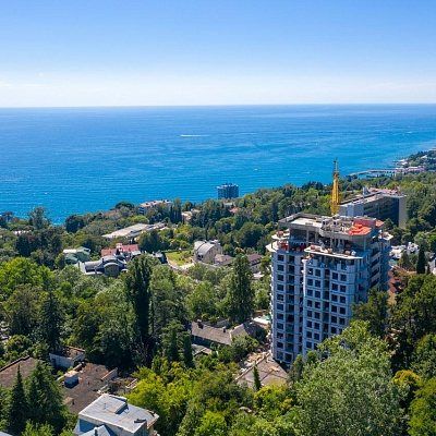 Строящийся дом с краном на крыше среди деревьев, за ним море до горизонта