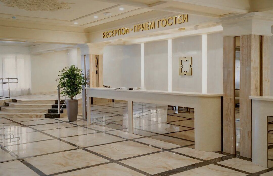 Лобби с мраморным полом, светлой стойкой приёма и надписью «Reception · Приём гостей» над ней, у лестницы — растение в кадке