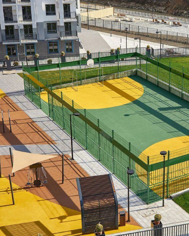 Спортивная площадка за сеткой с жёлтым и зелёным покрытием, рядом навесы и корпуса