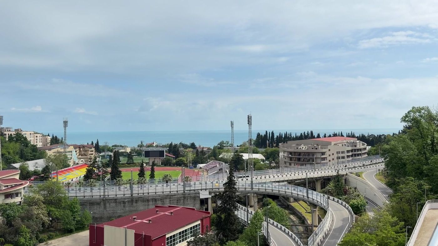 Вид с высокого этажа: городские кварталы, стадион с трибунами, эстакада дороги и море на горизонте