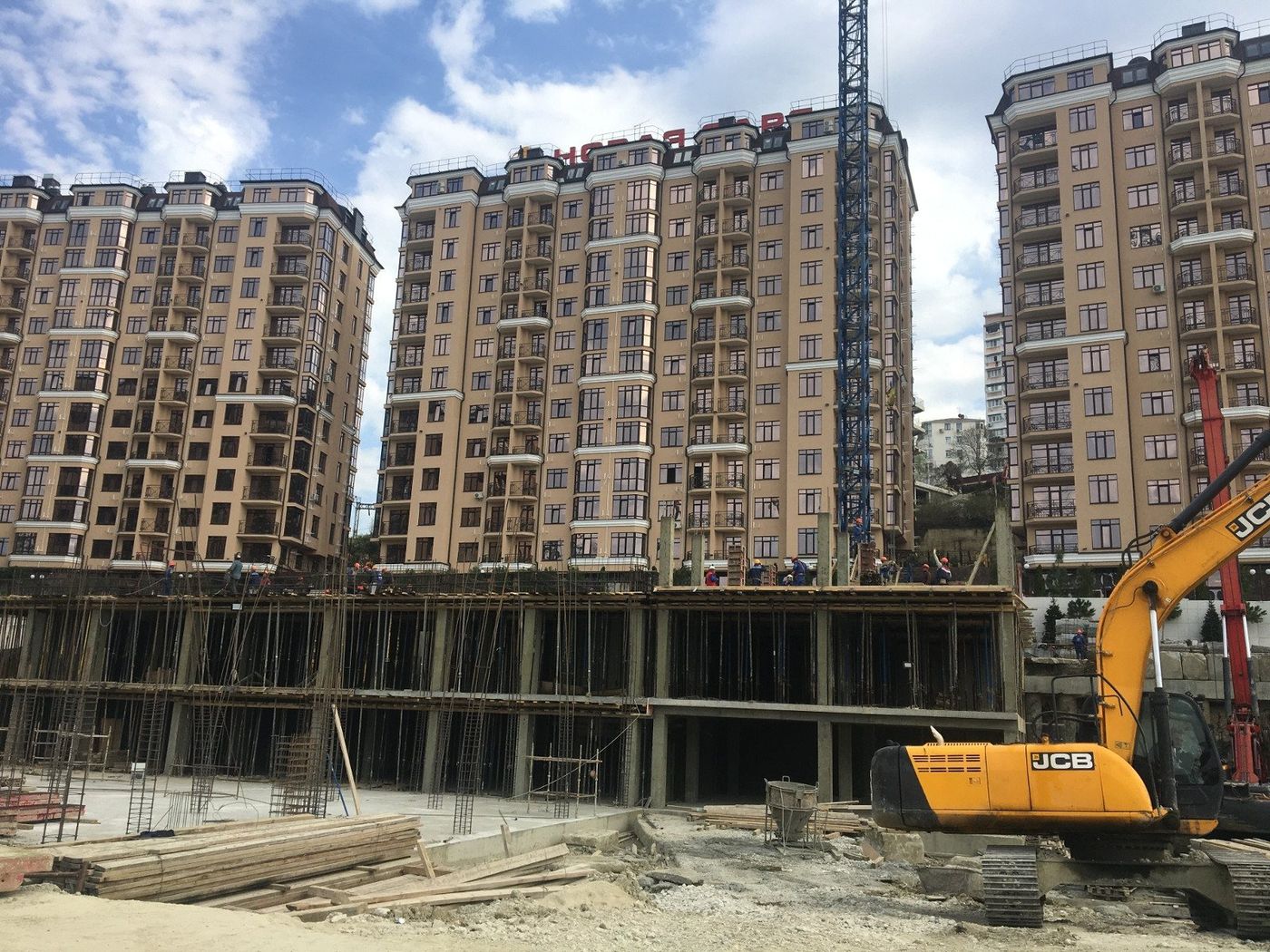 Три двенадцатиэтажных корпуса, перед ними стройплощадка с экскаватором