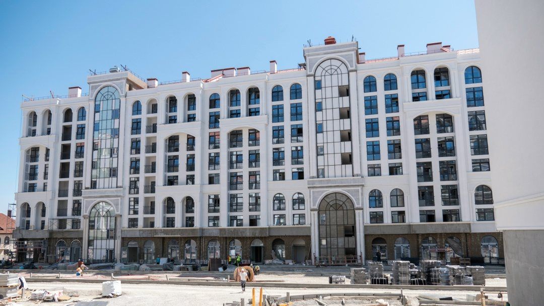 Девятиэтажный дом со светлым фасадом, арочными окнами и высоким остеклённым проёмом, перед ним строительная площадка